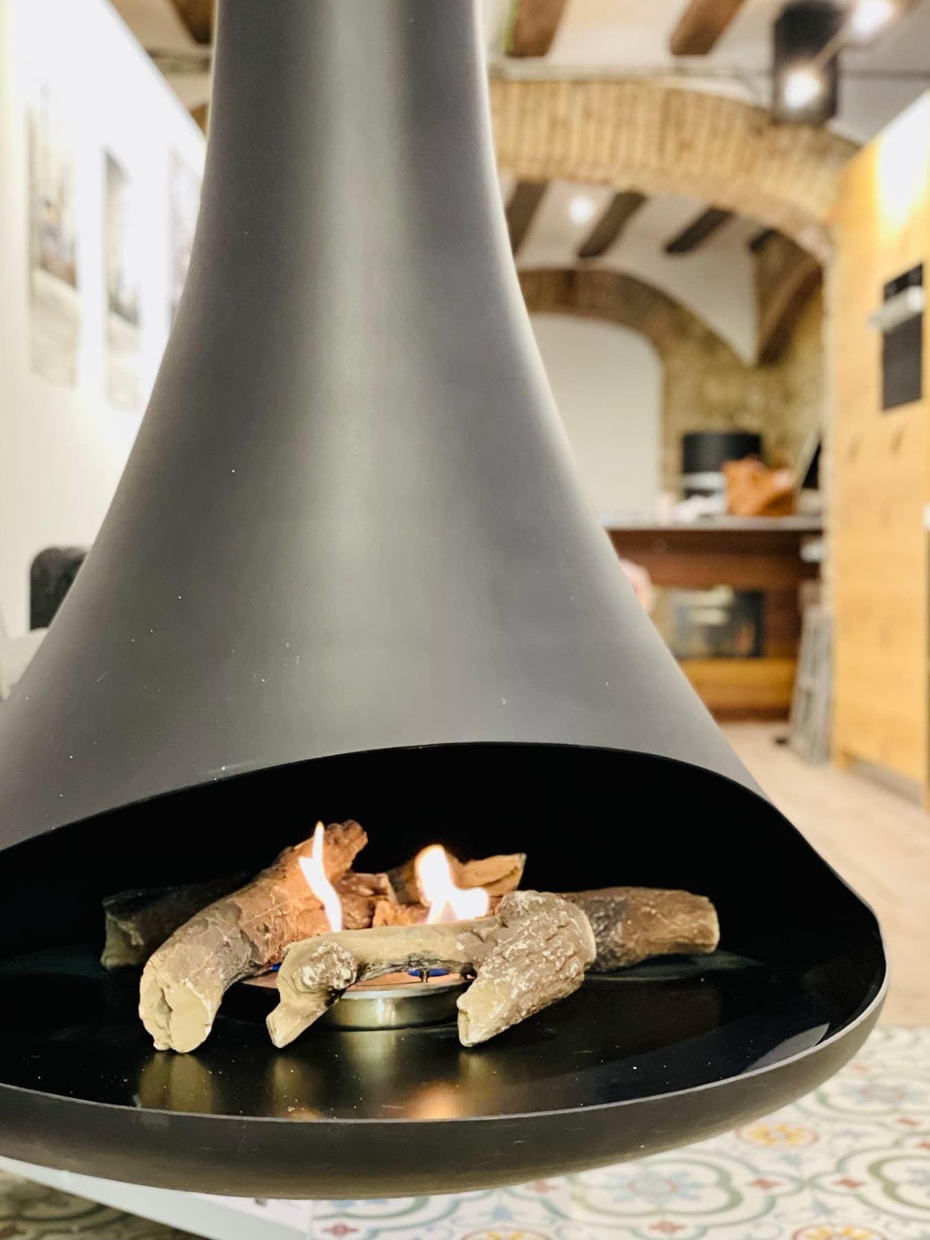 Suspended bioethanol fireplace with lit ceramic logs, under a ceiling of wooden beams and brick arches