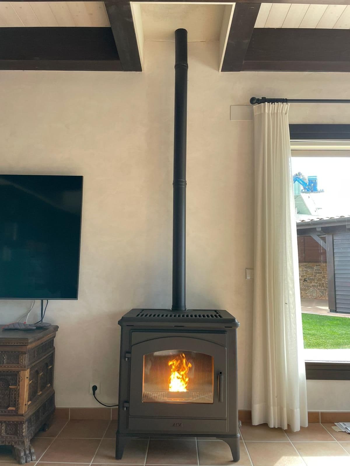Estufa de pellet negra encendida junto a una ventana, con el tubo de humos en vertical hasta el techo de madera